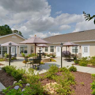 A well-maintained outdoor patio area with tables and umbrellas, surrounded by landscaping, adjacent to a building under a partly cloudy sky.