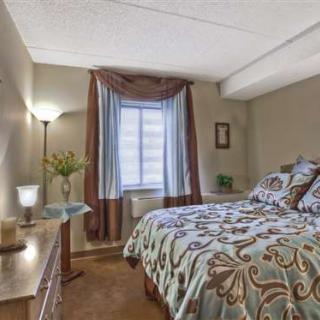 A cozy bedroom with a patterned bedspread, standing lamp, and natural light from a window with curtains.