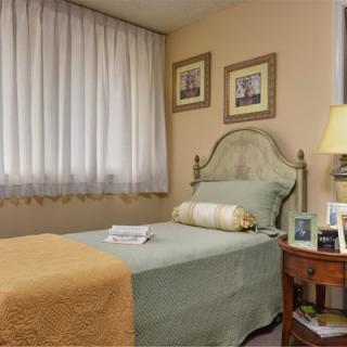A cozy bedroom featuring a single bed with green and orange bedding, complemented by a decorative pillow. A nightstand with a lamp and framed photos is beside the bed, while curtains softly filter the natural light.