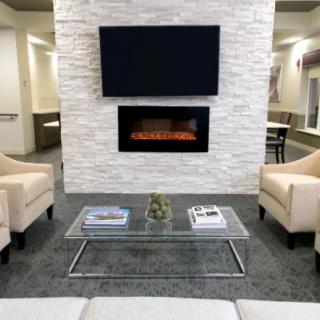 A modern living room with a mounted TV and fireplace, featuring a glass coffee table between two beige armchairs.