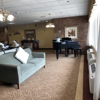 A cozy living room with a sofa and grand piano, featuring carpet flooring, brick walls, and large windows.