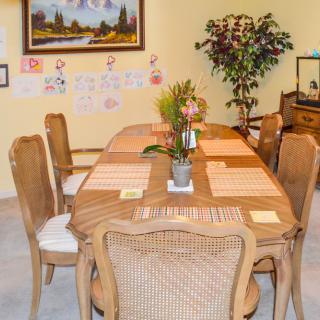 A cozy dining room with a wooden table set for six, adorned with placemats and a centerpiece, surrounded by chairs and decorated with wall art and plants.