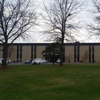 A large building with a parking lot, surrounded by trees and a grassy lawn under an overcast sky.