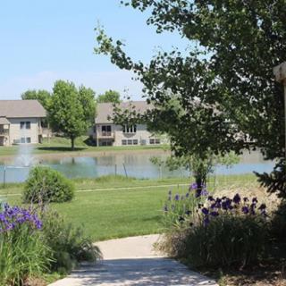 A scenic view of houses by a lake with a pathway, surrounded by trees and flowers.