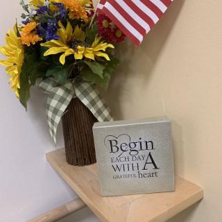 A decorative arrangement featuring a vase of colorful flowers, an American flag, and a sign with an inspirational quote on a small shelf.