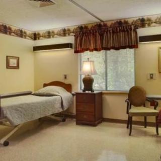 A hospital room with a single bed, a lamp on a wooden nightstand, and a chair near the window.