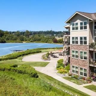A scenic view of an apartment building by a lakeside with a pathway and lush greenery under a clear blue sky.
