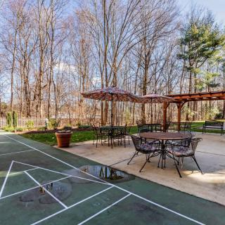 A backyard patio with a pergola and seating area next to a shuffleboard court surrounded by trees.