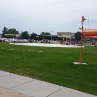 A spacious green field with a red flag, set against a cloudy sky and surrounded by a parking lot.