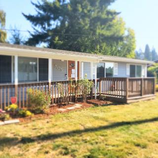A single-story house with a wooden deck and surrounding greenery on a sunny day.