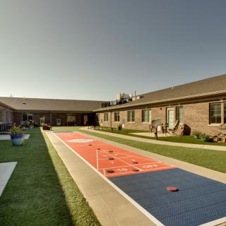 An outdoor shuffleboard court surrounded by brick buildings and a well-maintained lawn under clear skies.