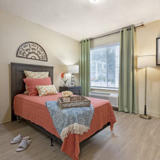 A cozy bedroom featuring a single bed adorned with decorative pillows and a colorful blanket, complemented by a lamp and window with green curtains.