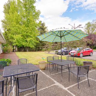 A cozy outdoor seating area with tables and umbrellas, surrounded by lush greenery and a parking lot.