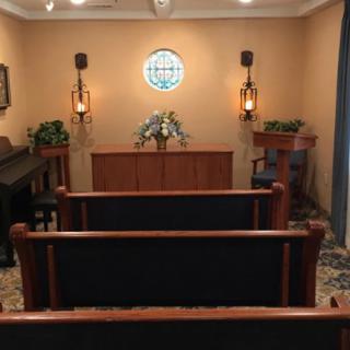 A small chapel interior with rows of wooden pews facing a modest altar, adorned with decorative sconces and floral arrangements.