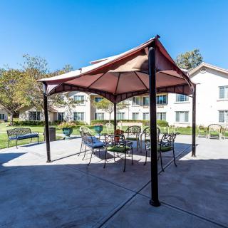A sunny outdoor area featuring a gazebo with patio furniture, surrounded by benches and greenery, with buildings in the background.