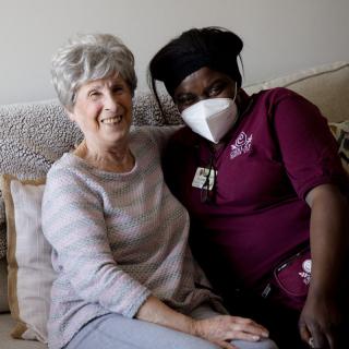 Two individuals sitting on a couch, one wearing a mask and a caregiver uniform.