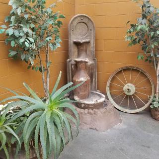 A rustic outdoor setup featuring a decorative fountain, potted plants, and an old wagon wheel against a warm-colored wall.