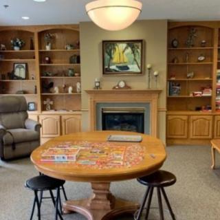 A cozy living room featuring a round table with a puzzle in progress, surrounded by bookshelves and an armchair near a fireplace.