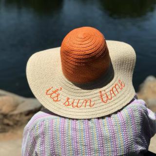 A person wearing a sun hat with the phrase 'it's sun time' embroidered on it, overlooking a lake.