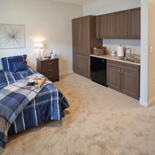 A cozy bedroom with a small kitchenette, featuring a bed with blue plaid bedding and wooden furniture.