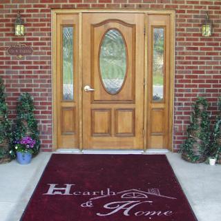 A welcoming wooden door with a decorative welcome mat on a brick porch, framed by potted plants.