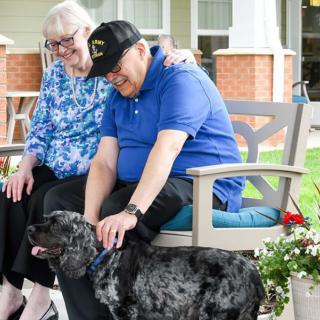 An elderly couple sitting on a bench outdoors, affectionately petting a dog.