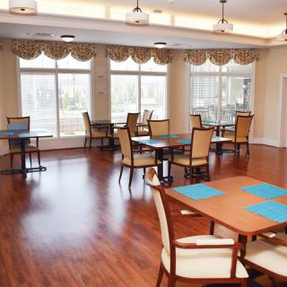 A bright and spacious dining room with hardwood floors, featuring several tables and chairs arranged neatly near large windows.