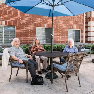 Three seniors sit around a table under a blue umbrella on a patio.