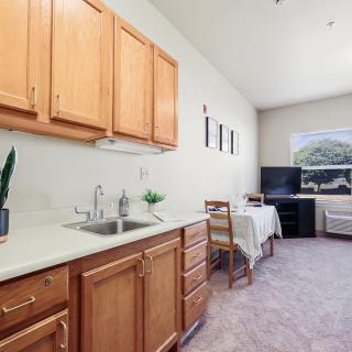 A compact kitchen area with wooden cabinets and a sink, leading to a small dining space with a table, chairs, and a television.