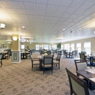 A spacious and well-lit dining area with multiple tables and chairs arranged on a carpeted floor, surrounded by large windows.