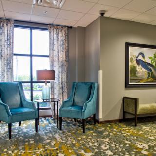 A cozy waiting area featuring two blue chairs, a small table with a lamp, and wall artwork.