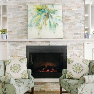 A cozy living room with a fireplace, flanked by two patterned chairs and a colorful artwork above.