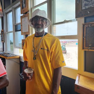 A man enjoying a drink while dressed in a vibrant yellow shirt and bucket hat, standing by the window.