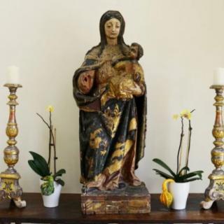 A wooden statue of the Virgin Mary holding baby Jesus, flanked by two ornate candles and potted orchids on an altar.