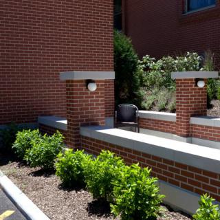 A brick wall with built-in lighting, surrounded by neatly landscaped plants and a bench.