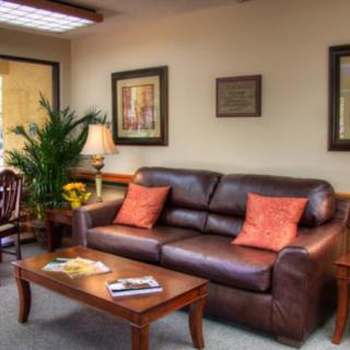 A cozy office waiting room featuring a leather sofa with orange pillows, artwork on the walls, a wooden coffee table, and a large potted plant.