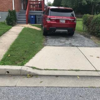 A red SUV parked in a driveway next to a sidewalk and lawn in front of a brick house.