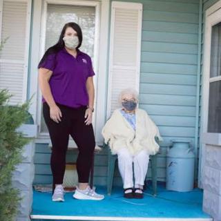 A caregiver stands beside an elderly person on a porch, both wearing masks.