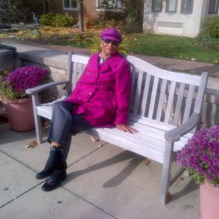 A person sitting on a bench wearing a purple coat and hat, surrounded by potted flowers.