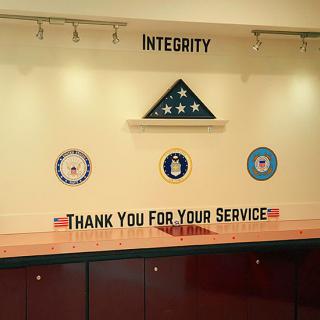 A tribute wall displaying a folded American flag and military emblems with the words 'Thank You For Your Service' and 'Integrity'.