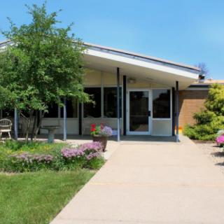 A small building with a pathway leading to its entrance, surrounded by green landscaping and trees under a clear blue sky.