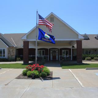 A building with American and state flags at the entrance, surrounded by a flower bed and parking area on a sunny day.