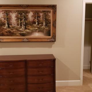 A bedroom interior featuring a wooden dresser, a framed painting of a forest scene, and an open closet with neutral-colored walls.