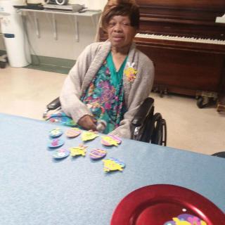 An elderly woman sitting in a wheelchair indoors, with decorations on the table and a piano in the background.