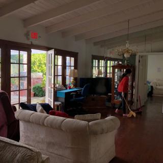 A cozy living room with large windows and open doors letting in natural light, featuring a sofa and television, while someone cleans the wooden floor.