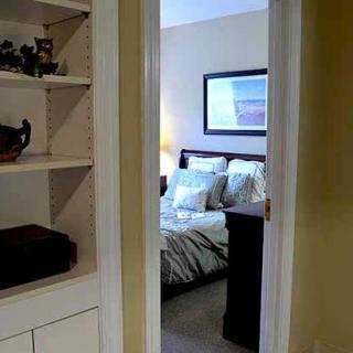 A view into a cozy bedroom through an open doorway, with a neatly arranged bed and a built-in shelf on the side.