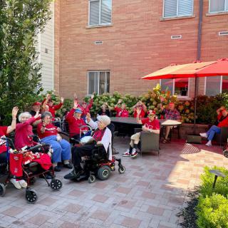 A group of elderly individuals enjoying a sunny day outdoors, gathered under a red umbrella.