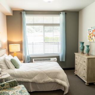 A cozy and well-lit bedroom with a neatly made bed, a lamp on a side table, and a dresser adorned with decorative items.