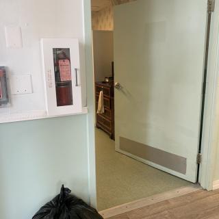 An open door leads from a hallway with a fire extinguisher and a garbage bag to an interior room.