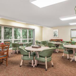 A well-lit community dining area featuring multiple tables and green chairs, surrounded by large windows.